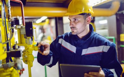 Serious dedicated plant worker in working clothes and with protective helmet on head screwing valve and holding tablet.